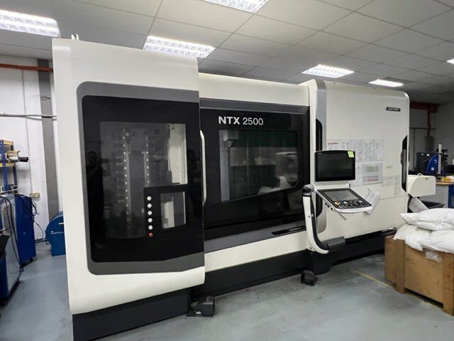 A white NTX 2500 CNC machine with a control panel is situated in an industrial workshop setting under fluorescent lighting.